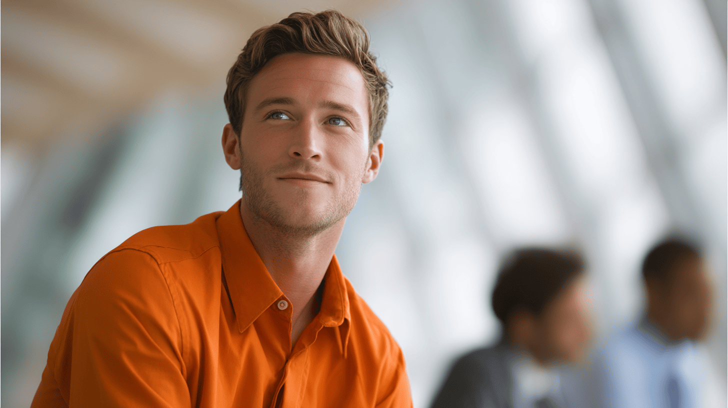 A young man in an orange shirt smiles while looking thoughtfully off-camera, with blurred figures in the background.