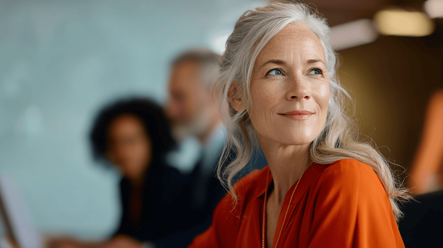 A woman with long, grey hair and an orange blouse looks thoughtfully while seated in a modern office setting.