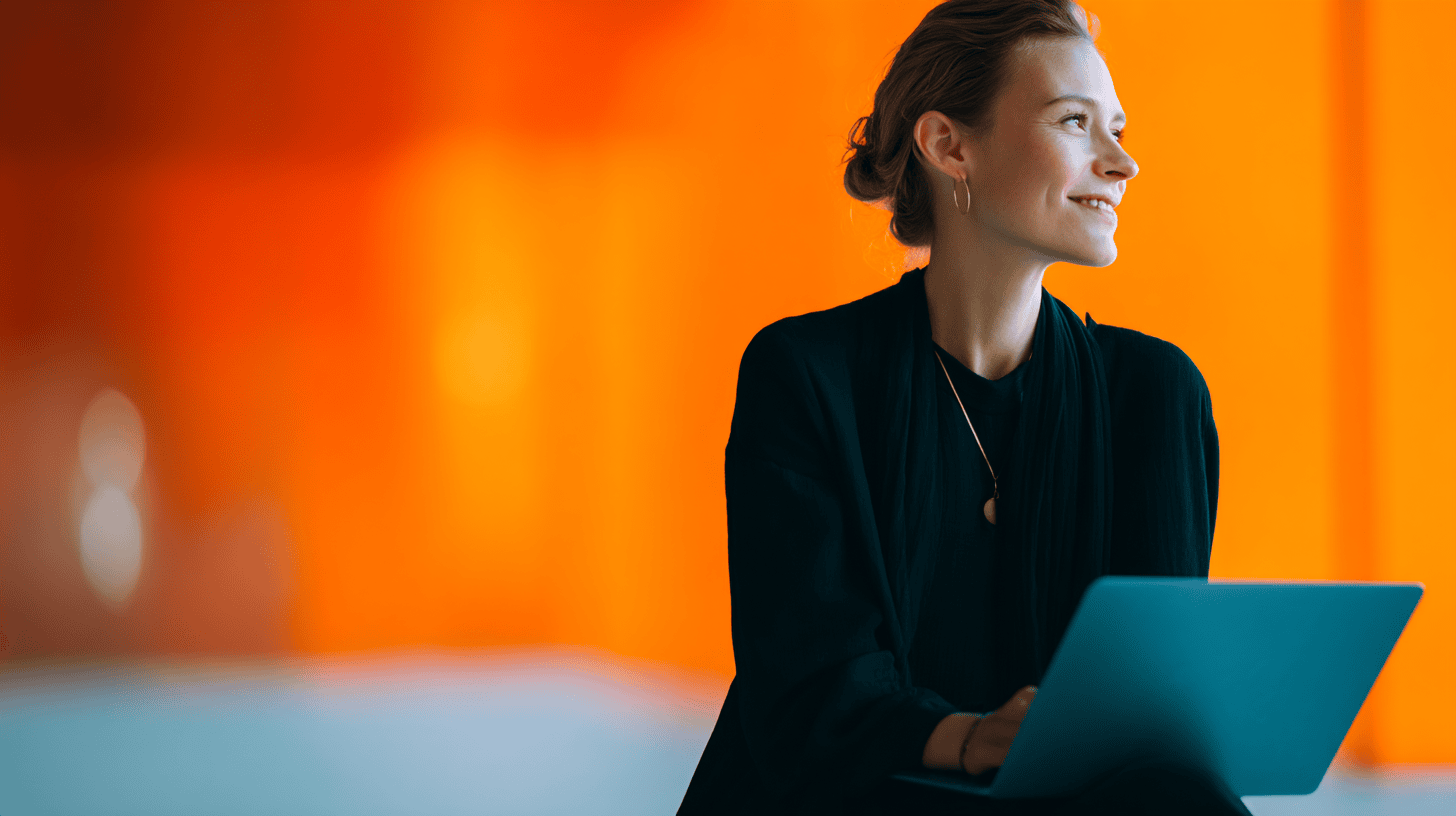 A woman in a black outfit sits smiling, looking at a laptop against a vibrant orange background.