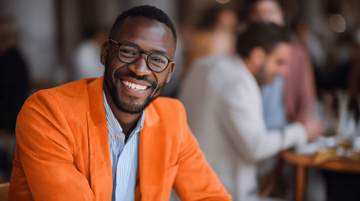 Smiling man in an orange jacket, glasses, seated at a table in a busy cafe. Blurry background with people and drinks.