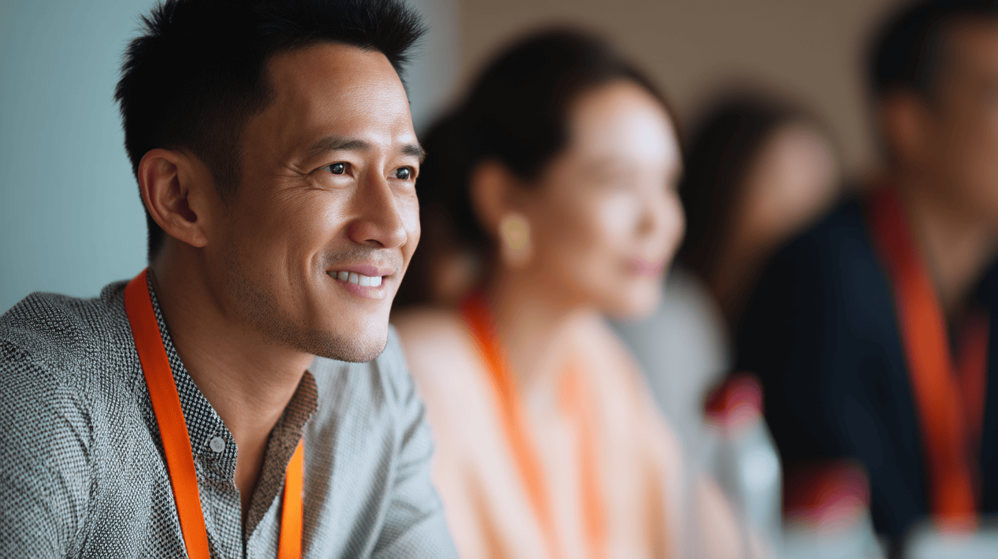 A smiling man in a patterned shirt and orange lanyard sits at a conference table, with blurred attendees behind him.