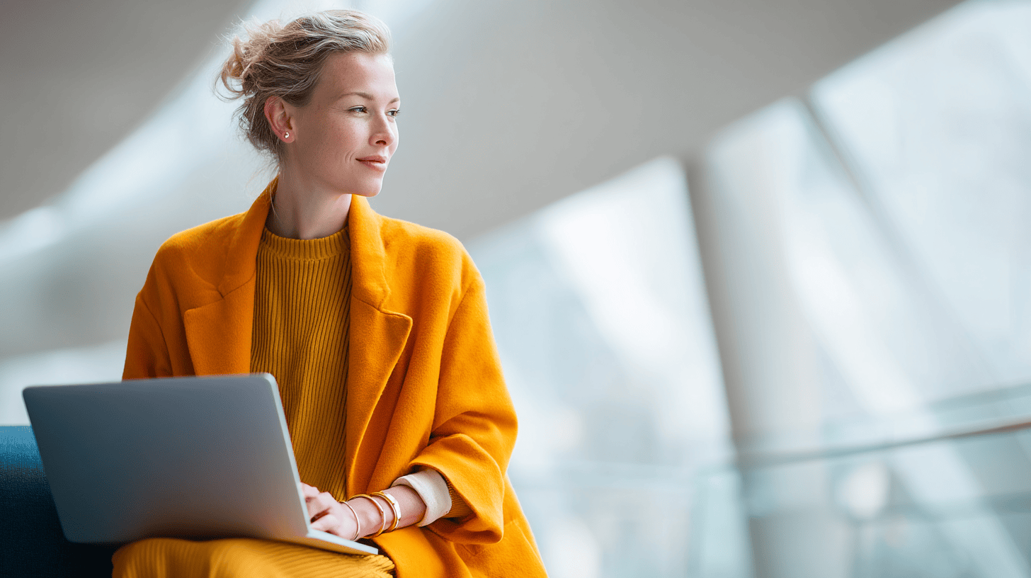 A woman in a bright orange coat and sweater sits with a laptop, looking thoughtfully out a large window.
