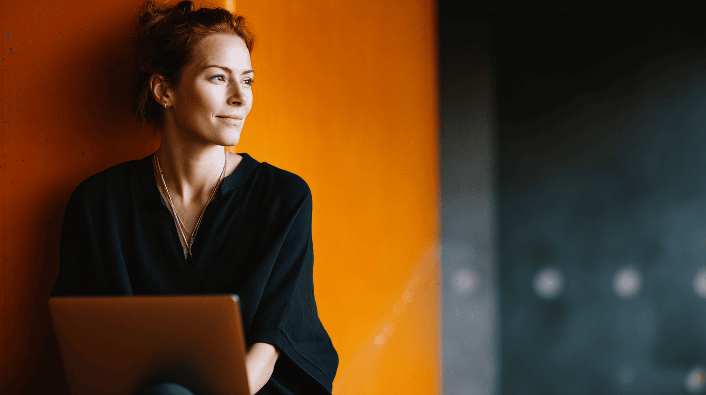 A woman in a black blouse sits against an orange wall, holding a laptop with a contemplative expression.