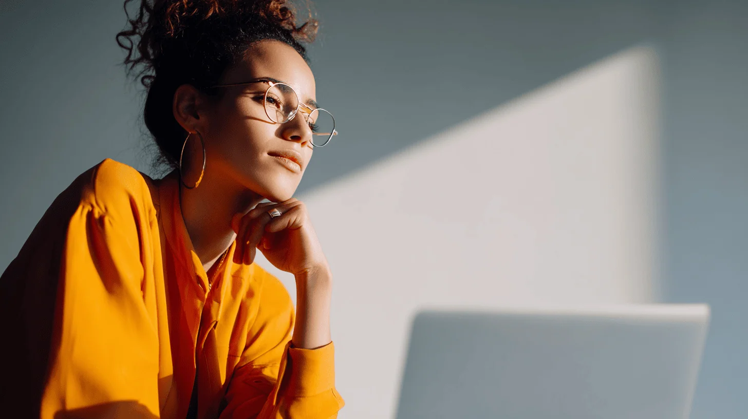 A woman in an orange shirt and round glasses sits thoughtfully beside a laptop, with sunlight creating a shadow on a wall.