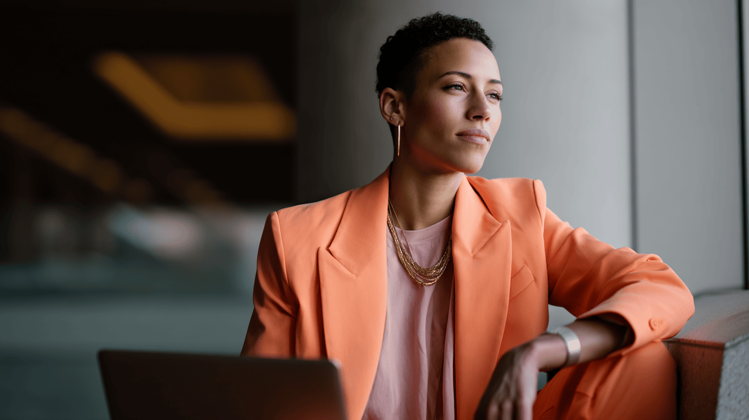 A woman in an orange blazer sits beside a laptop, gazing thoughtfully out of a modern, brightly lit space.