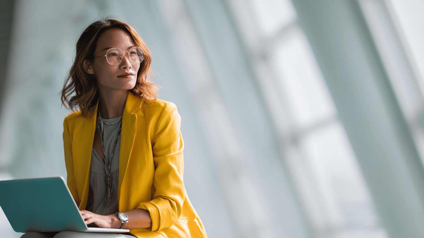A woman in a yellow jacket sits with a laptop, looking thoughtfully in a bright, modern workspace.