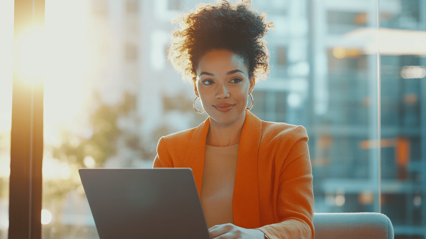 A woman in an orange blazer smiles while working on a laptop, with bright sunlight and a blurred city backdrop.