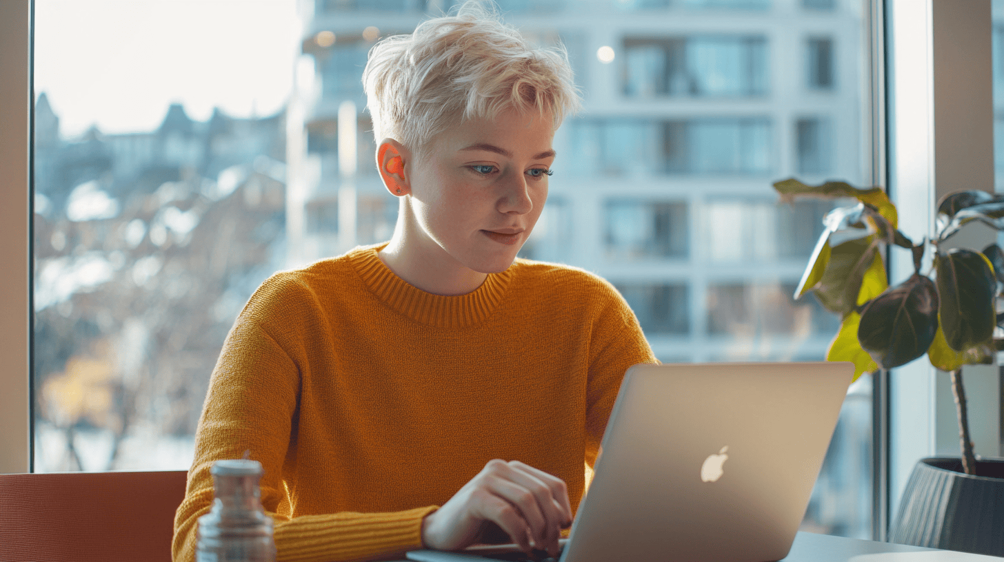 A young person in a yellow sweater works on a laptop by a window, with a plant nearby and a cityscape in the background.