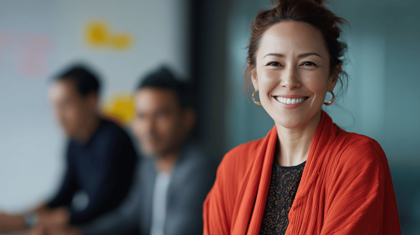 A smiling woman in an orange scarf sits in a meeting with two blurred individuals in the background. Bright, modern setting.