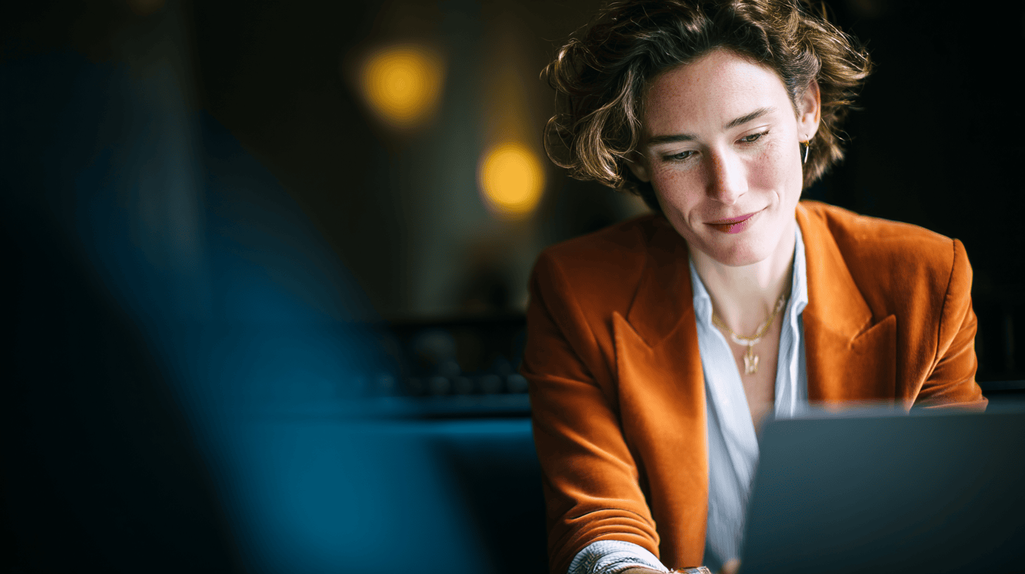 A woman with curly hair is smiling while working on a laptop in a cozy, dimly lit setting. She wears an orange blazer.