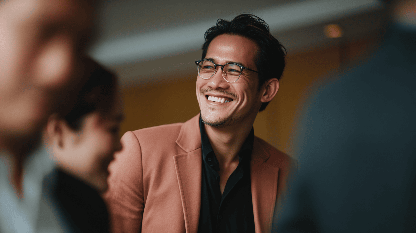 Smiling man in a pink blazer and glasses, engaging in conversation with blurred figures in the foreground.