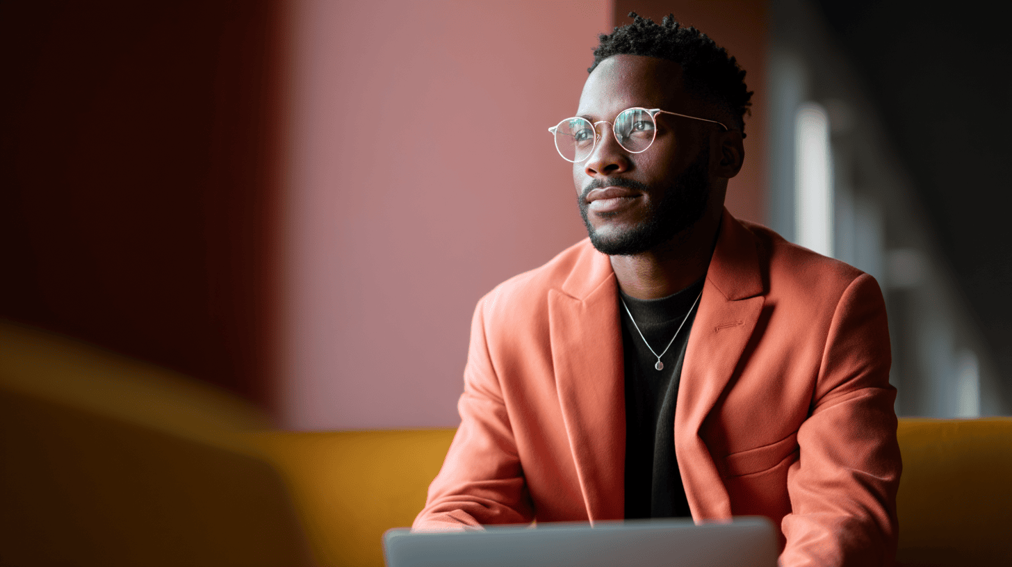A young man with round eyeglasses sits at a laptop in a softly lit modern space. He wears a coral-orange blazer over a black shirt and a thin pendant necklace, gazing thoughtfully out of frame toward a nearby window.